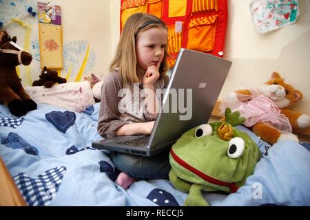 Fille, 7 ans, en travaillant avec un ordinateur à la maison dans sa chambre, assise sur son lit superposé, faire ses devoirs pour l'école Banque D'Images