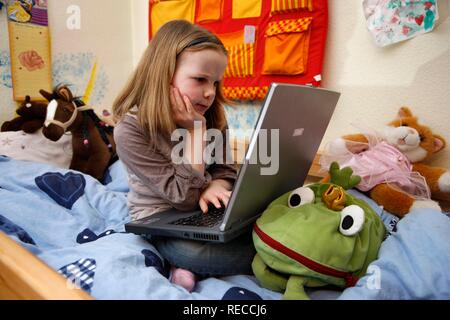 Fille, 7 ans, en travaillant avec un ordinateur à la maison dans sa chambre, assise sur son lit superposé, faire ses devoirs pour l'école Banque D'Images