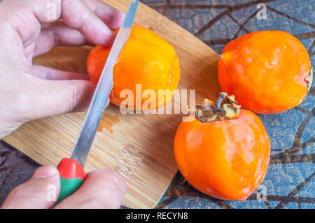 Vue du dessus de la main la coupe d'un kaki des fruits sur un plaque à découper. D'autres fruits sont conservés sur la table Banque D'Images