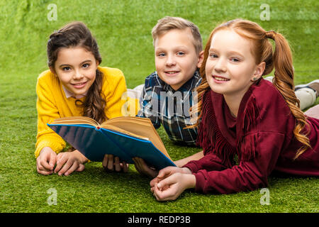 Cute preteen schoolkids allongé sur pelouse verte, holding book and looking at camera Banque D'Images