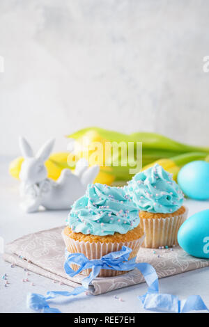 Cupcakes à la vanille décoré d'asperge et ruban bleu. Bunny, les oeufs colorés et de tulipes jaunes à l'arrière-plan. Banque D'Images