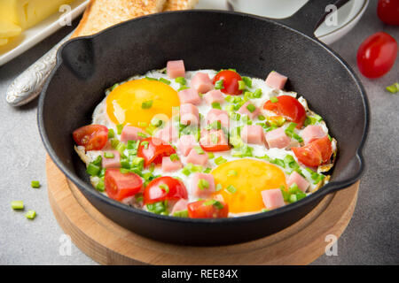 Oeufs frits en noir poêle avec du jambon, tomates cerises et oignons verts de printemps. Délicieux petit déjeuner avec du pain grillé et du beurre. Le classique Anglais Banque D'Images