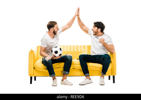 Happy young man sitting on couch with soccer ball et offrant un rapport cinq isolated on white Banque D'Images