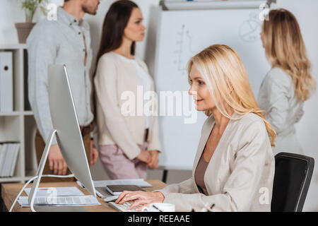 Vue latérale du smiling businesswoman en utilisant l'ordinateur de bureau et les jeunes collègues travaillant avec tableau blanc derrière Banque D'Images