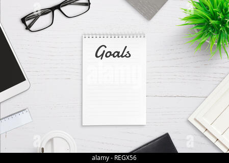 Buts d'écriture sur le bloc-notes vide avec la page de titre dans l'en-tête. Objectifs Vue de dessus d'un bureau avec téléphone intelligent, tasse de café, des verres, des végétaux. Banque D'Images