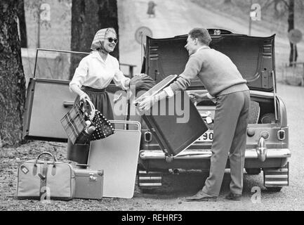 Couple dans les années 60. Un jeune couple d'emballage sont le coffre de leur voiture avec les bagages et matériel de camping pour une location de vacances. Une table pliante pratique est aussi emballé. Elle est habillée dans un manoir typique des années 50 avec une chemise blanche, jupe large, lunettes de soleil et ses cheveux dans un foulard. Suède 1957 Banque D'Images