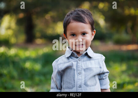 Un mignon petit garçon mexicain debout dans un domaine boisé vert, avec sourire doux sur son visage, et portant une chemise à manches longues. Banque D'Images