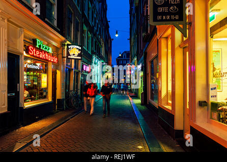 Les gens sur la rue étroite passant par allumé en magasins, restaurants et bars dans le vieux quartier historique d'Amsterdam, Pays-Bas. Banque D'Images