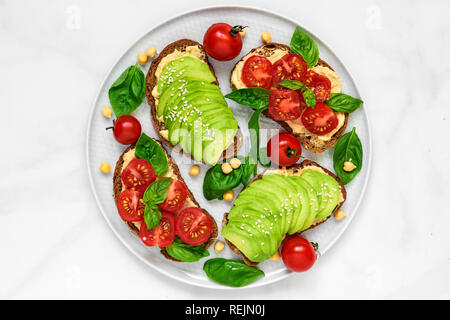 L'avocat et les tomates toasts avec du hoummos, sésame et basilic dans une plaque sur fond de marbre blanc. La nourriture végane. Vue de dessus Banque D'Images