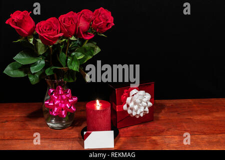 Belles roses rouges dans un vase avec noeud rose wrapped present et bougie rouge avec étiquette-nom sur la table en bois sur fond sombre. La Saint-Valentin, fête des mères, de l'évaluation environnementale Banque D'Images