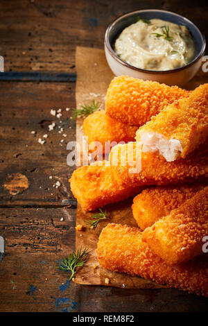 Les bâtonnets de poisson croustillants ou des bâtons dans la poêle ou au four à la chapelure servi sur du papier brun sur une table rustique avec une sauce tartare Banque D'Images