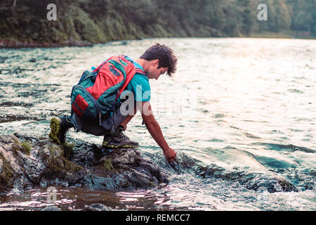 Jeune garçon avec sac à dos, assis sur un rocher surplombant une rivière, tenant la main dans l'eau, à l'eau, il porte des vêtements de sport Banque D'Images
