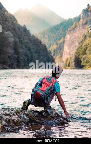 Jeune garçon avec sac à dos, assis sur un rocher surplombant une rivière, tenant la main dans l'eau, à la montagne, à pics, il porte des vêtements d'été sports Banque D'Images