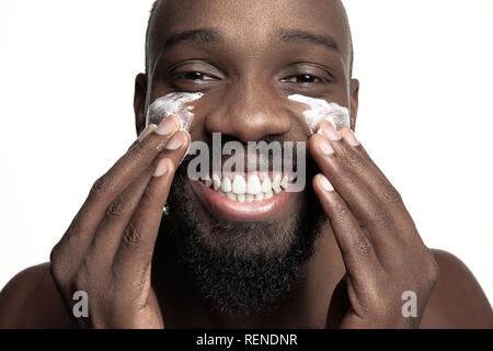 Les jeunes africains-américains guy l'application de la crème pour le visage sous ses yeux sur fond blanc. Portrait of a young happy smiling african man au studio. La haute couture modèle masculin. Banque D'Images