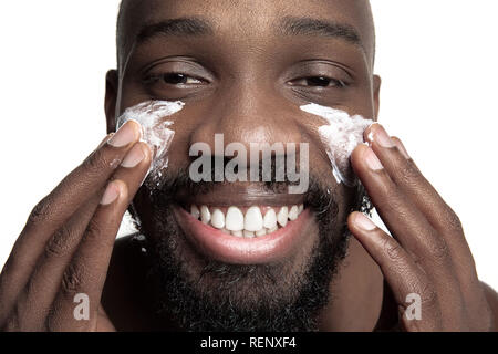 Les jeunes africains-américains guy l'application de la crème pour le visage sous ses yeux sur fond blanc. Portrait of a young happy smiling african man au studio. La haute couture modèle masculin. Banque D'Images