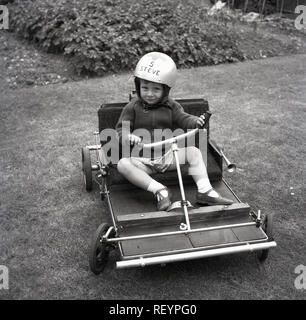 1967, historiques, jeune garçon dans un jardin à l'arrière assis dans un go-kart, portant son casque personnalisé, England, UK. La construction d'un bricolage a go-kart ou passe-temps populaire pour beaucoup de projet à cette époque. Banque D'Images