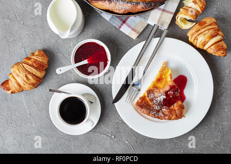 Une partie de baby néerlandais garnie de confiture de framboises sur une plaque avec fourchette et couteau, croissants, tasse de café et de crème fraîche sur une table, la vue Banque D'Images