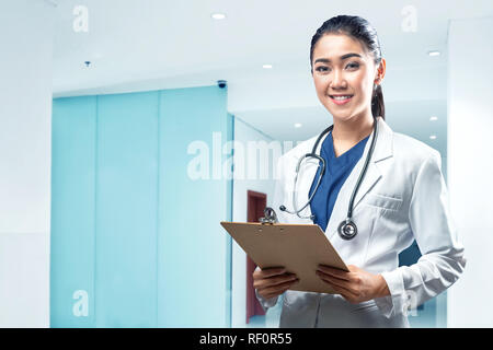 Jolie femme asiatique médecin vêtu de blanc manteau avec stéthoscope et presse-papiers sur l'hôpital Banque D'Images