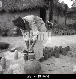 Années 1950, historiques, de vêtements traditionnels africains locaux Yoruba hat, un Aso Oke hat et le chiffon robe, faire des pots d'argile à l'extérieur de sa hutte, le Nigeria, l'Afrique. Banque D'Images