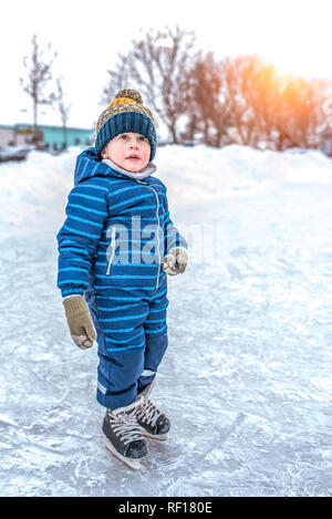 Un petit garçon de 4-7 ans est de patinage. Premiers pas sur des patins. En hiver dans la ville sur la patinoire. Dans un chapeau chaud et combinaison bleue. Banque D'Images