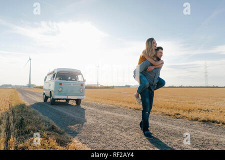 Young man carrying girlfriend piggyback on dirt track à camper van in rural landscape Banque D'Images