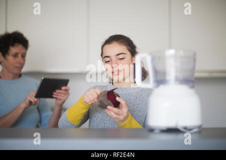 Portrait of smiling young woman peeling betterave rouge dans la cuisine pendant que maman à l'aide de tablette dans l'arrière-plan Banque D'Images