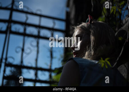 Vue de côté peur girl wearing headband debout derrière la porte de hauteur au crépuscule. Jolie jeune femme avec des cheveux blonds bouclés perdu dans parc de nuit. Mystères cachés par l'obscurité Banque D'Images