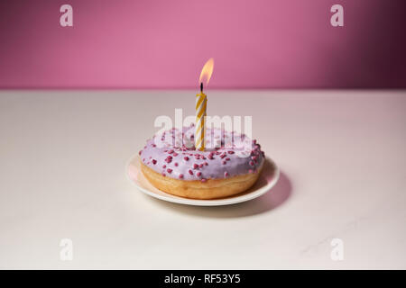 Bougie brûlante au milieu de donut avec sprinkles sur table blanche sur fond rose Banque D'Images