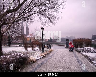 Une femme dans un manteau rouge et d'une poussette rouge dans un parc sur un jour nuageux avec chute de neige et de neige au sol avec vue sur Ostrow Tumski dans le background Banque D'Images