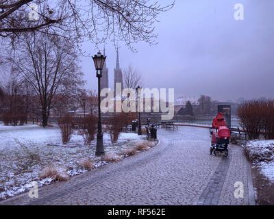 Une femme dans un manteau rouge et d'une poussette rouge dans un parc sur un jour nuageux avec chute de neige et de neige au sol avec vue sur Ostrow Tumski dans le background Banque D'Images