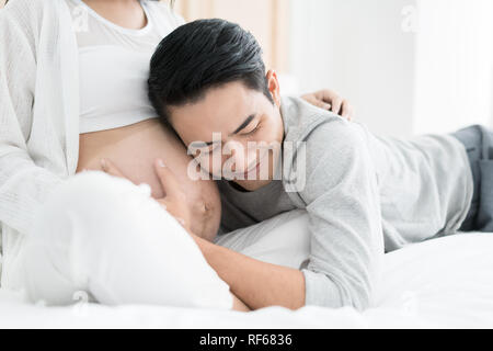 Bel homme asiatique est à l'écoute de sa belle épouse asiatique enceinte le ventre et sourit. Famille amour concept. Banque D'Images