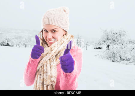 Femme vêtue de rose pullover avec foulard et hat showing Thumbs up comme geste portant des gants violet comme en hiver la neige fond paysage Banque D'Images