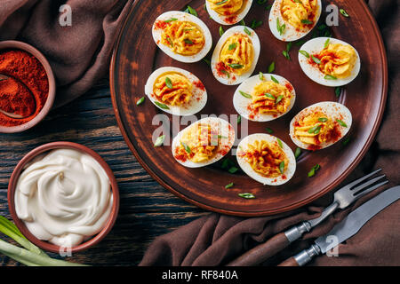 Oeufs farcis saupoudré de paprika et de l'oignon vert haché finement sur une plaque de faïence sur une vieille table en bois rustique avec tissu brun, vue d'un Banque D'Images