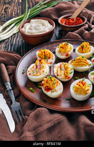 Oeufs farcis saupoudré de paprika et de l'oignon vert haché finement sur une plaque d'argile sur une vieille table rustique avec un tissu marron, verres à Mayo et paprik Banque D'Images