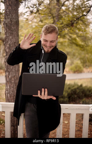 Un jeune souriant et happy man holding ordinateur portable à la main, saluant quelqu'un avec un geste (main levée) par vidéo conférence téléphonique, pourrait être commu Banque D'Images