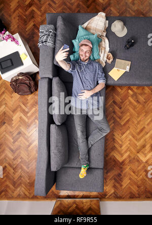 Un jeune homme souriant, parlant avec son selfies smart-phone, pose sous un canapé. Vue de dessus de toute sa chambre, pleine longueur a tourné. Banque D'Images