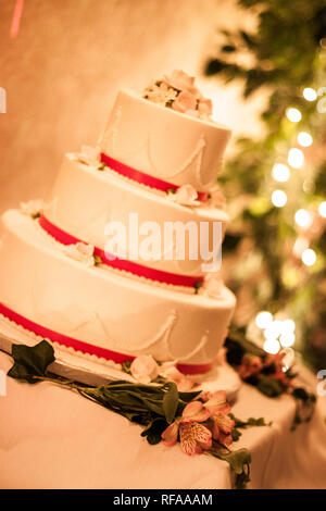 Gâteau de mariage à l'écran aux côtés de fleurs, sur une table, avec feux lumineux en arrière-plan, à une réception de mariage. Banque D'Images