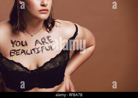 Cropped shot of woman surdimensionnée en soutien-gorge noir. Femme avec "Vous êtes belles", écrit sur son corps contre fond brun Banque D'Images