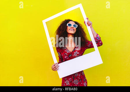 Jolie femme dans une robe rouge et des lunettes de se tenir contre un mur jaune et tenant un grand cadre vide. Beau modèle féminin posant avec pi vide Banque D'Images