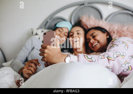 Girls lying on bed s'amusant de prendre un. selfies Trois jeunes filles bénéficiant d'un film sur téléphone mobile pendant une soirée pyjama en position couchée sur le lit. Banque D'Images