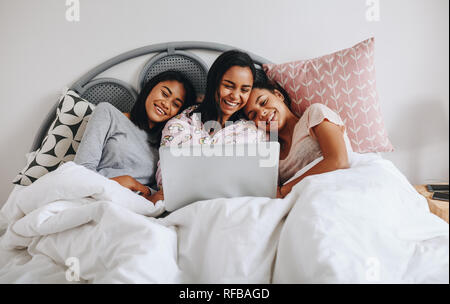 Girl friends ayant un temps heureux pendant une soirée pyjama de regarder un film sur ordinateur portable. Smiling girls sitting on bed with laptop jouissant de sa soirée pyjama. Banque D'Images