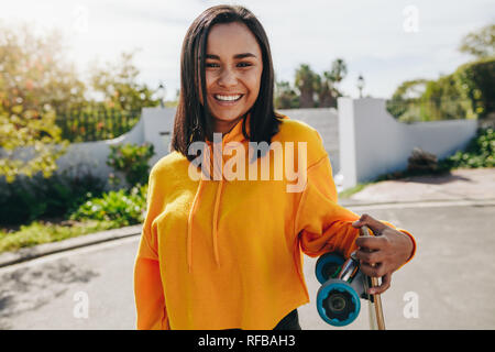 Cheerful woman standing on street tenant une planche à roulettes. Smiling Girl standing en plein air dans la rue sur une journée ensoleillée. Banque D'Images