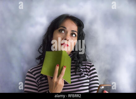 Jeune femme brune avec pullover rayé avec un livre sur son menton de penser à une question, isolé sur un fond gris Banque D'Images