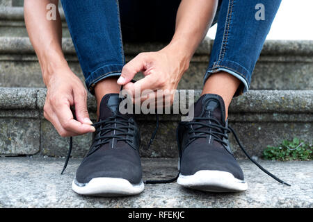 Libre d'un jeune homme de race blanche, vêtu d'un pantalon en jean, assis sur un escalier dans la rue, lier ou délier les lacets de ses baskets Banque D'Images