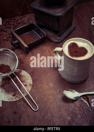 Photo d'une table en bois rustique, de grains de café, d'une meuleuse et une tasse de café avec du chocolat en forme de cœur saupoudrer. Banque D'Images