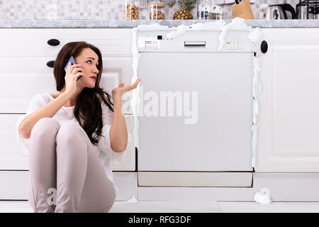 Triste femme appelant Technicien sur téléphone cellulaire pour réparer lave-vaisselle avec mousse venant de C Banque D'Images