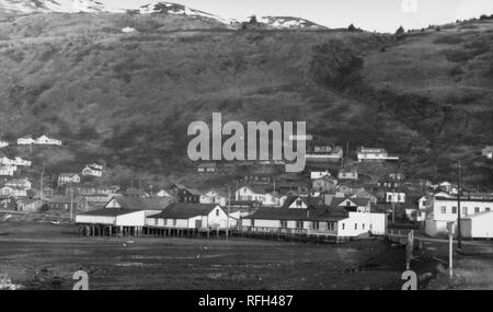 Photographie en noir et blanc de Kodiak, avec un fond marin à marée basse au premier plan, le 'O'Kraft et fils' general store visible sur une jetée, et plus de bâtiments en bois et les pointes des montagnes enneigées en arrière-plan, photographié au cours d'un voyage de chasse, de pêche situé dans l'Alaska, USA, 1955. () Banque D'Images