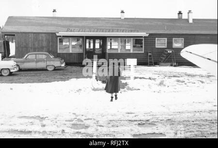 Photographie en noir et blanc d'une femme, tourné par derrière, en vue pleine longueur, portant une longue robe sombre, garni de fourrure et des bottes, marchant sur sol couvert de neige vers la porte ouverte d'un sol en bois, un seul niveau terminal de l'aéroport marqué 'Pacific Northern Airlines, ' avec les ailes d'un avion visible à droite au premier plan, photographié au cours d'un voyage de chasse, de pêche situé dans l'Alaska, 1955. () Banque D'Images