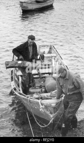 Photographie en noir et blanc de deux hommes d'âge moyen, un homme au premier plan à la bas comme il les plans tremblotants la proue d'un canot qui est tiré de la rive, une deuxième, brun, portant un sac de fusil, navigue la coque afin de parvenir à l'avant, avec de l'eau et d'un canot ancré visible à l'arrière-plan, photographié au cours d'un voyage de chasse, de pêche situé dans l'Alaska, 1955. () Banque D'Images