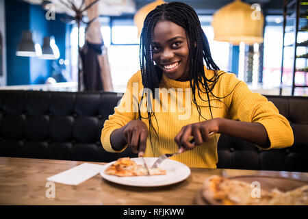 Les jeunes afro american woman wearing dans chandail jaune eating pizza in cafe Banque D'Images